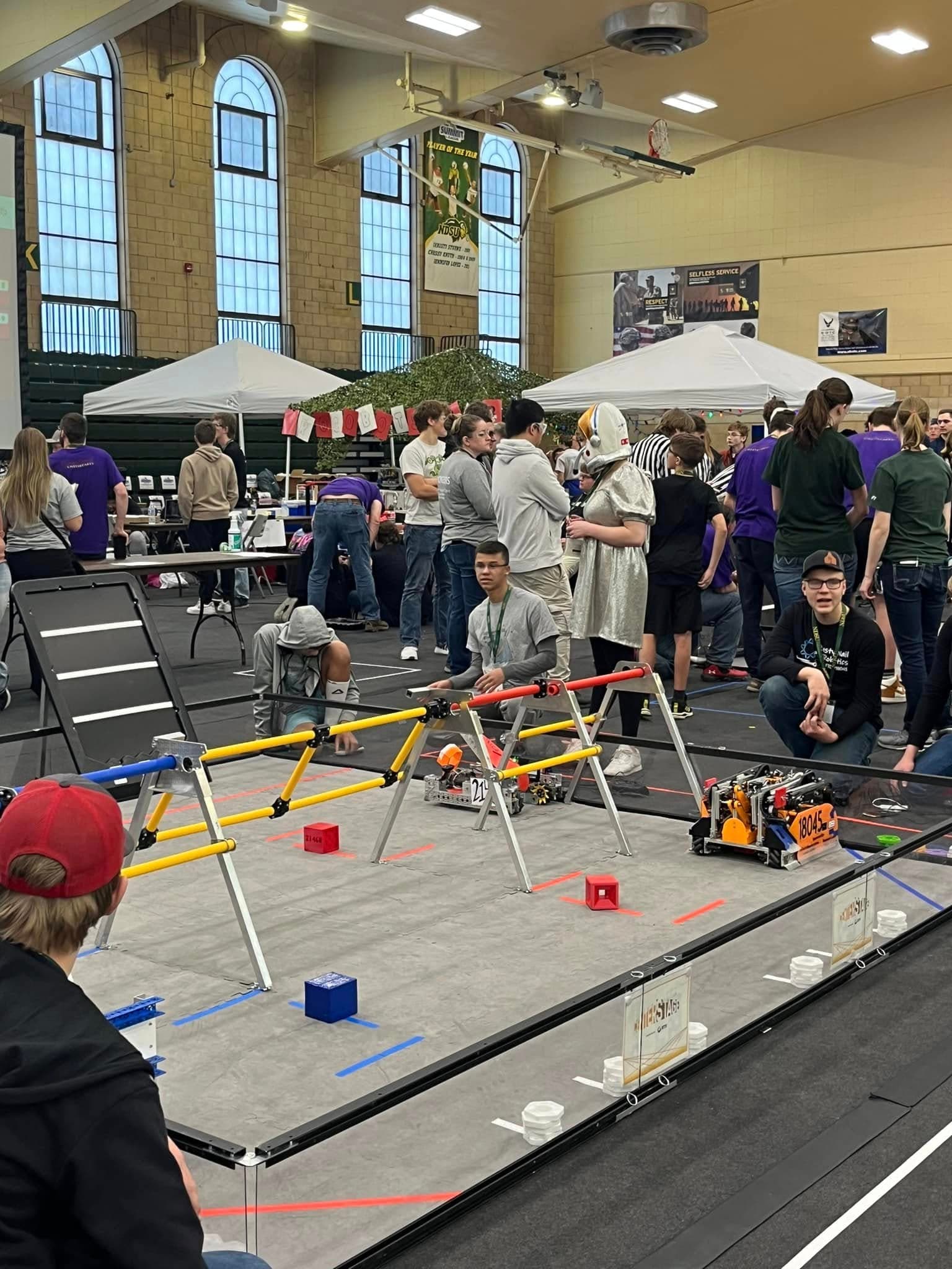FTC competition match — robots driving on the field, students and referees nearby.